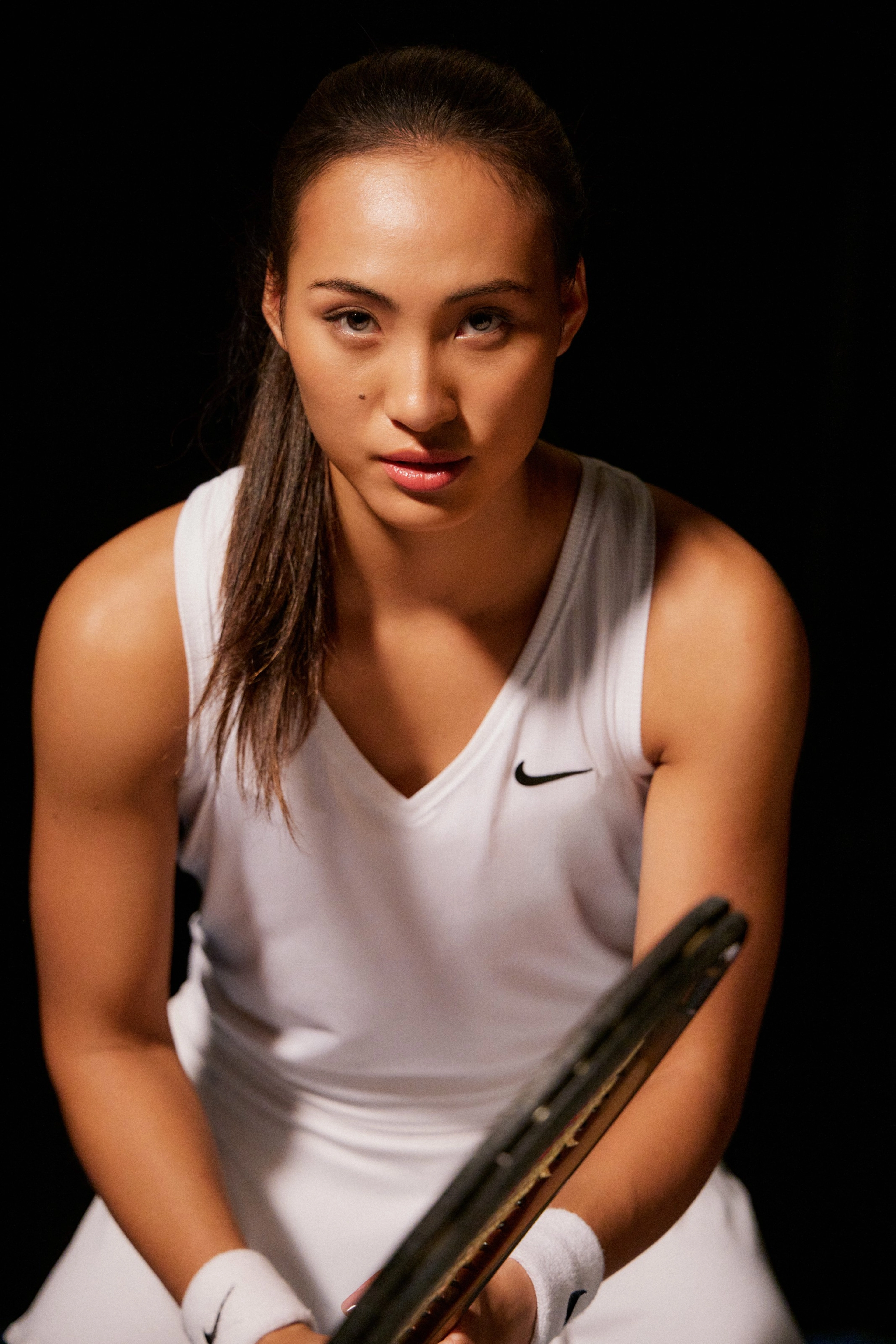 Front view of Qinwen Zheng directly facing the camera, while she holds her tennis racquet in front of her, prepared to receive a serve. She wears a white NikeCourt Victory tank with a black Swoosh, white tennis skirt, and white wristbands. She stands against a black background.