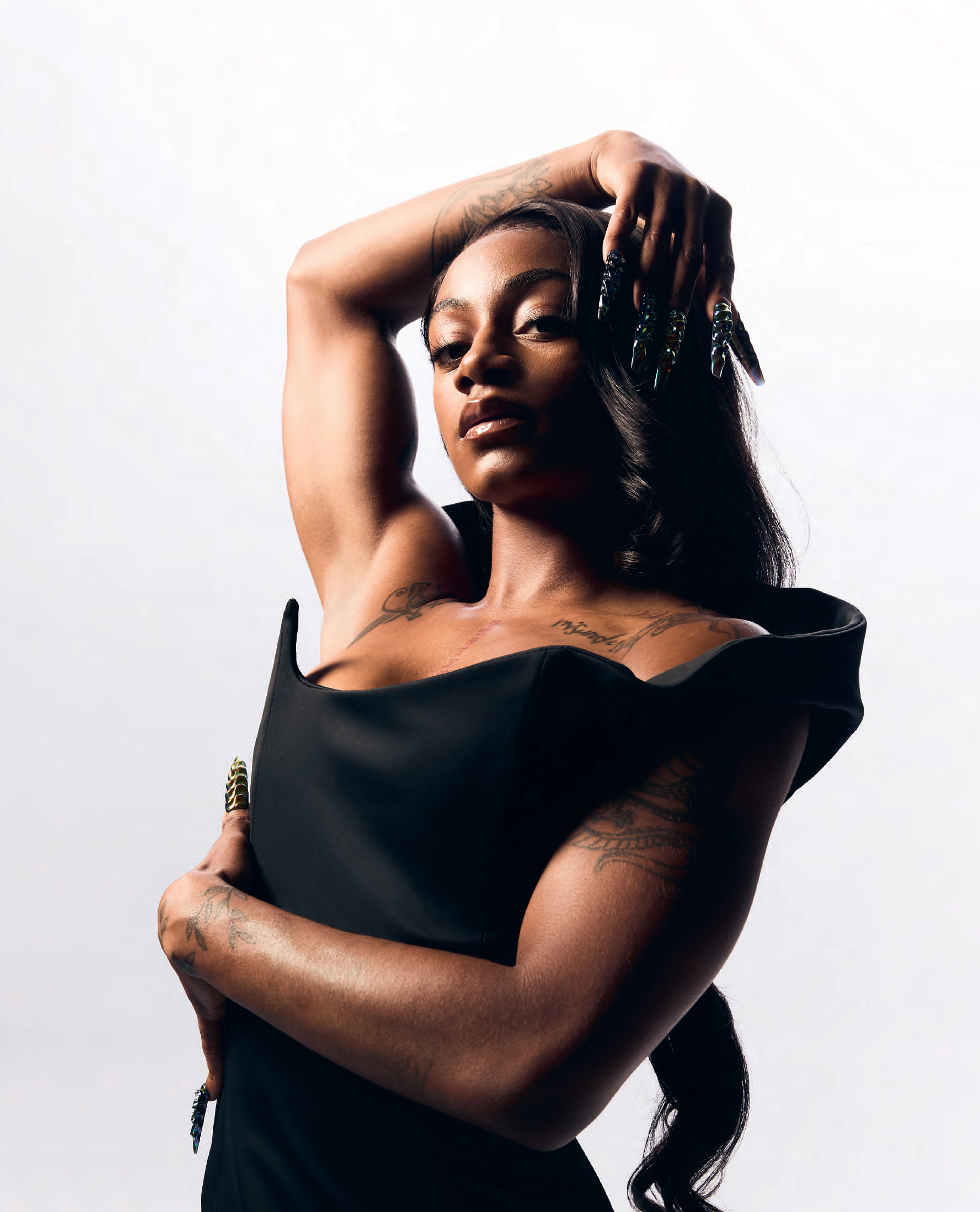 ShaCarri Richardson looks directly at the camera as she raises her arm over her head and crosses her body with her other arm. She wears a black dress, and we see her stylish nail art and tattoos.
