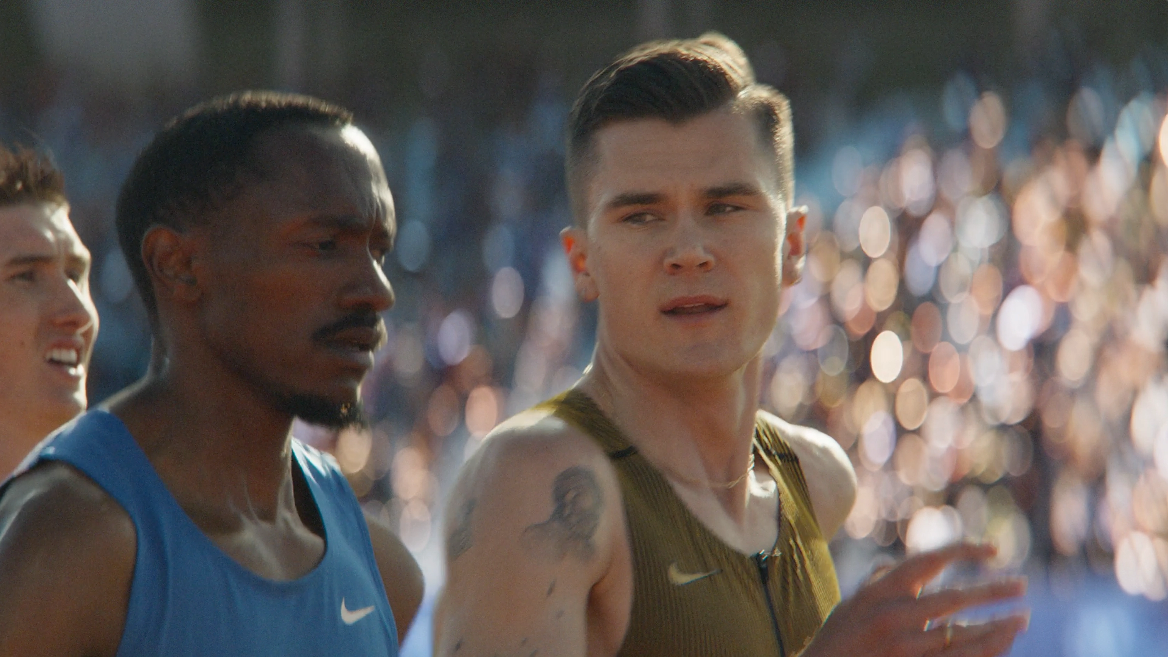Close up of Jacob Ingebrigtsen at a track event, alongside another competitor.  Jacob wears a gold Nike running singlet and there is a crowd in the background.