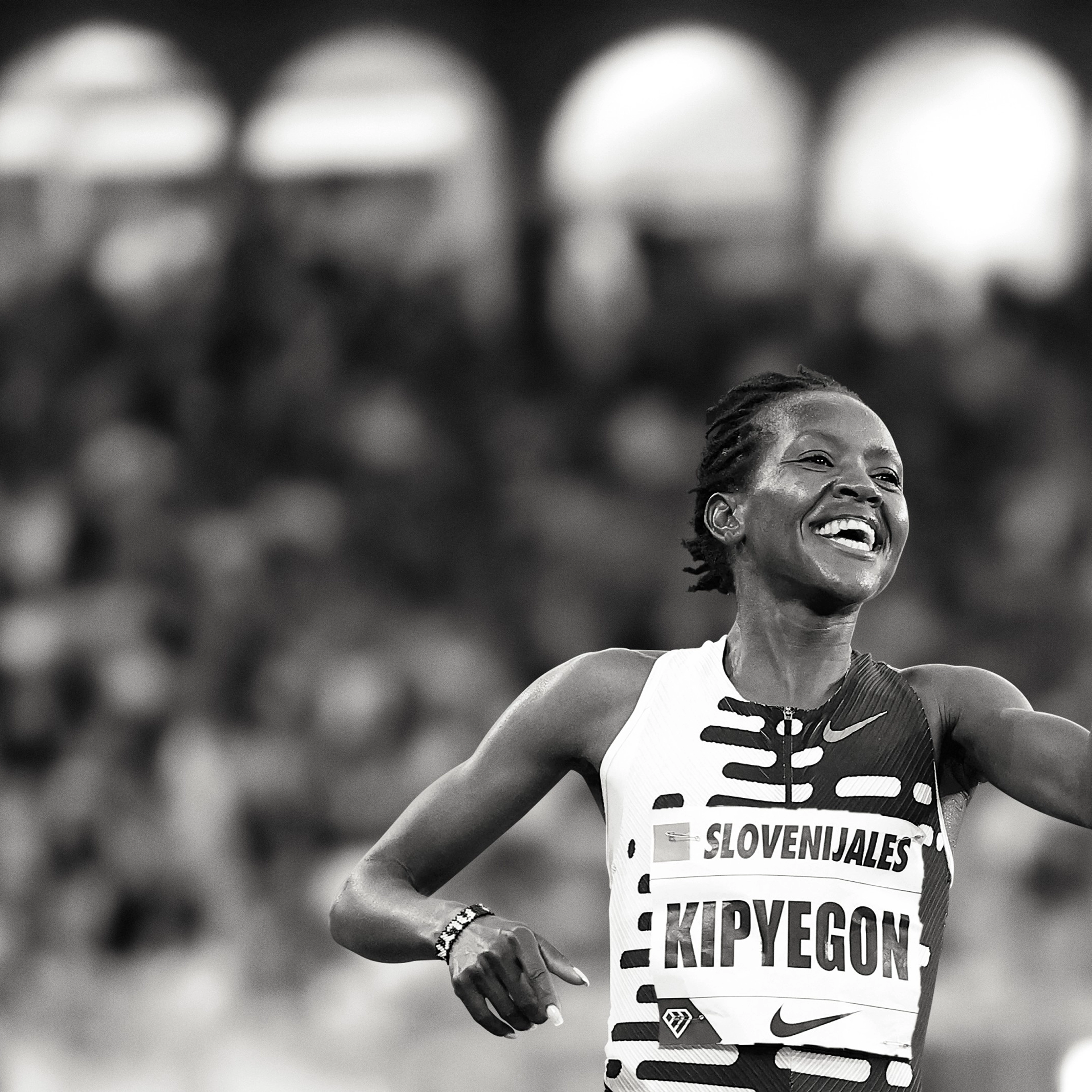 Black and white photo of Faith Kipyegon with a celebration smile and pointing a finger forward. She wears a patterned Nike running top, and there is a crowd behind her.