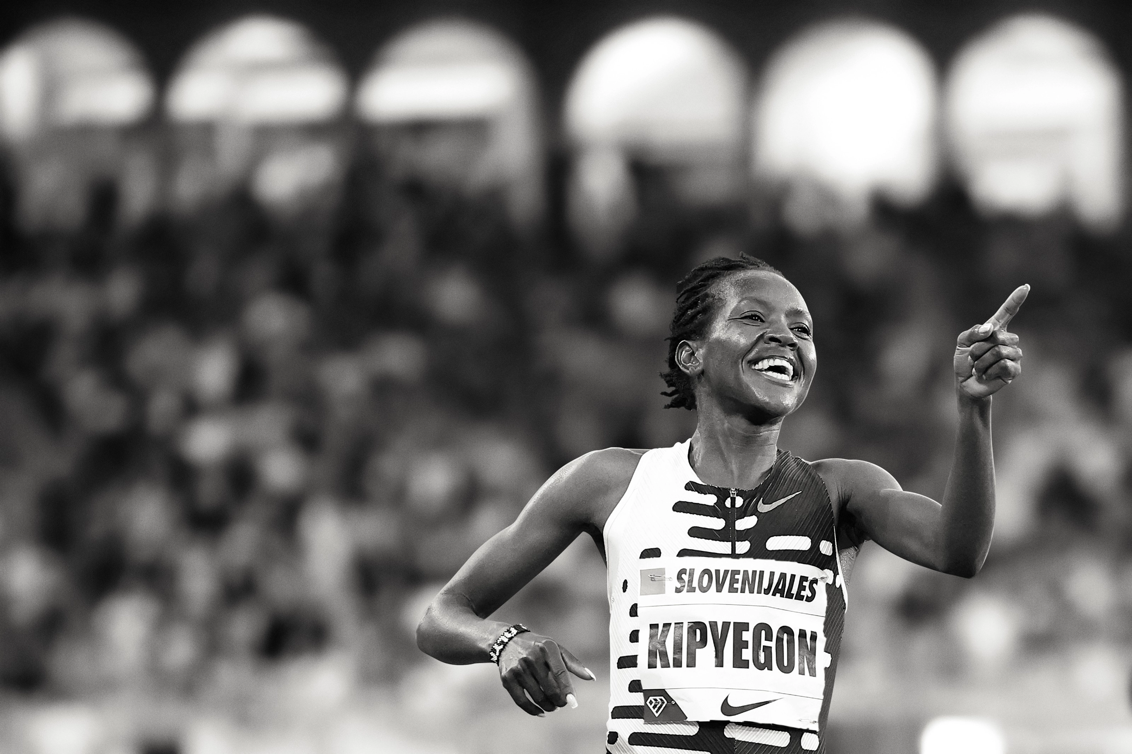Black and white photo of Faith Kipyegon with a celebration smile and pointing a finger forward. She wears a patterned Nike running top, and there is a crowd behind her.