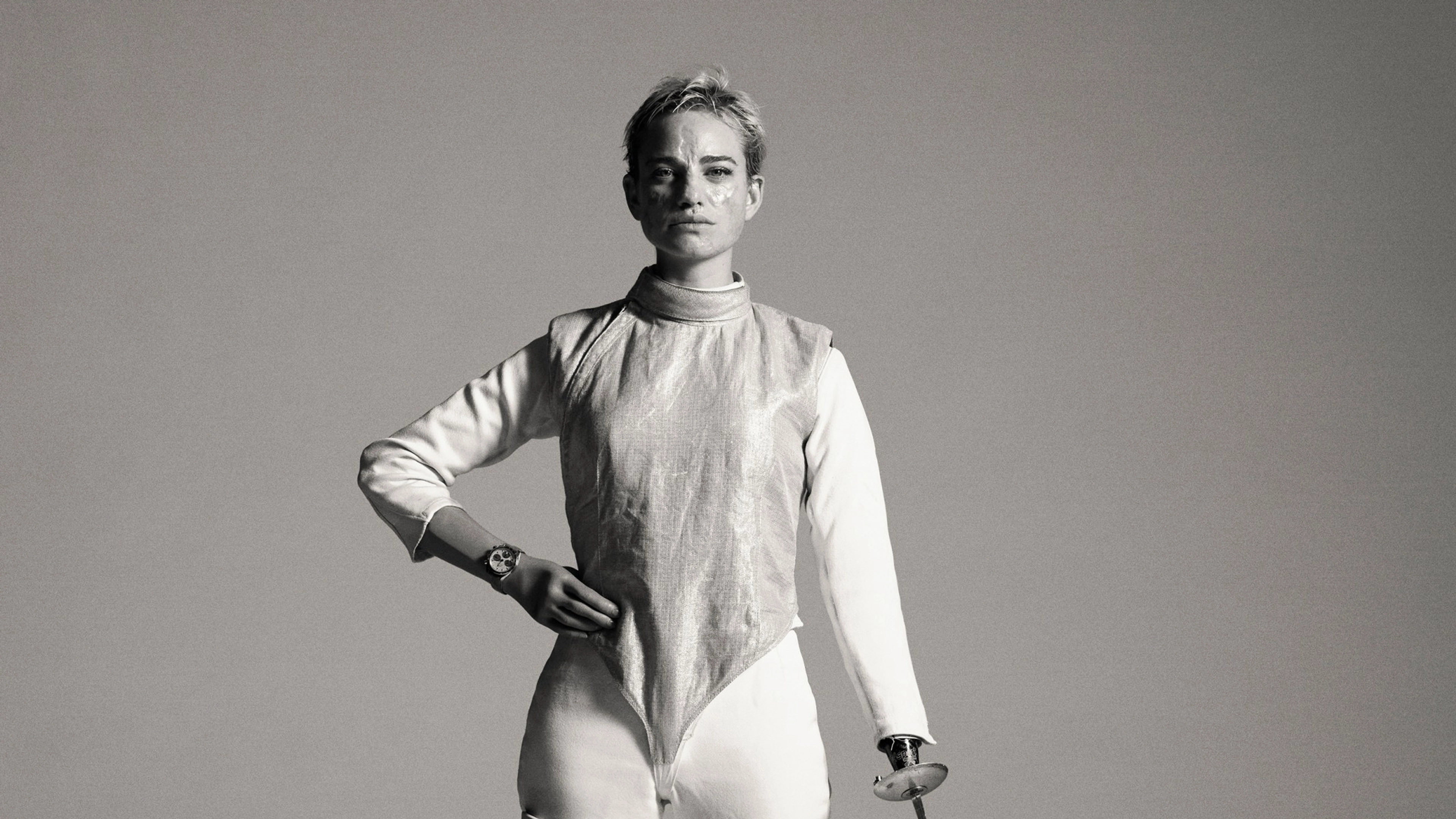 Black and white full body portrait of Bebe Vio, holding one hand to her hip and using her other arm to touch her fencing foil tip to the ground. Bebe looks unsmiling directly at the camera, as she stands in her fencing suit, standing on two prosthetic legs.