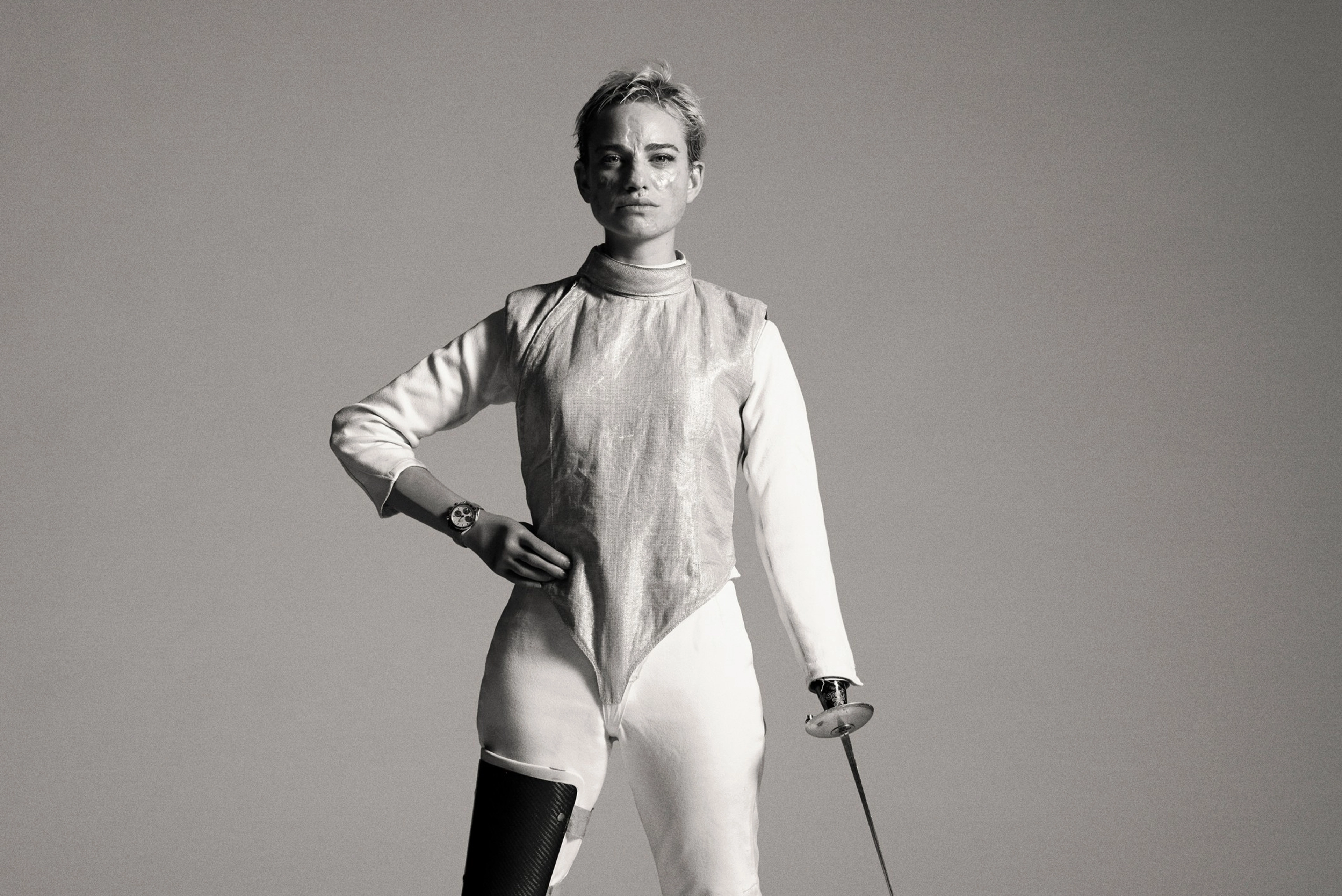 Black and white full body portrait of Bebe Vio, holding one hand to her hip and using her other arm to touch her fencing foil tip to the ground. Bebe looks unsmiling directly at the camera, as she stands in her fencing suit, standing on two prosthetic legs.