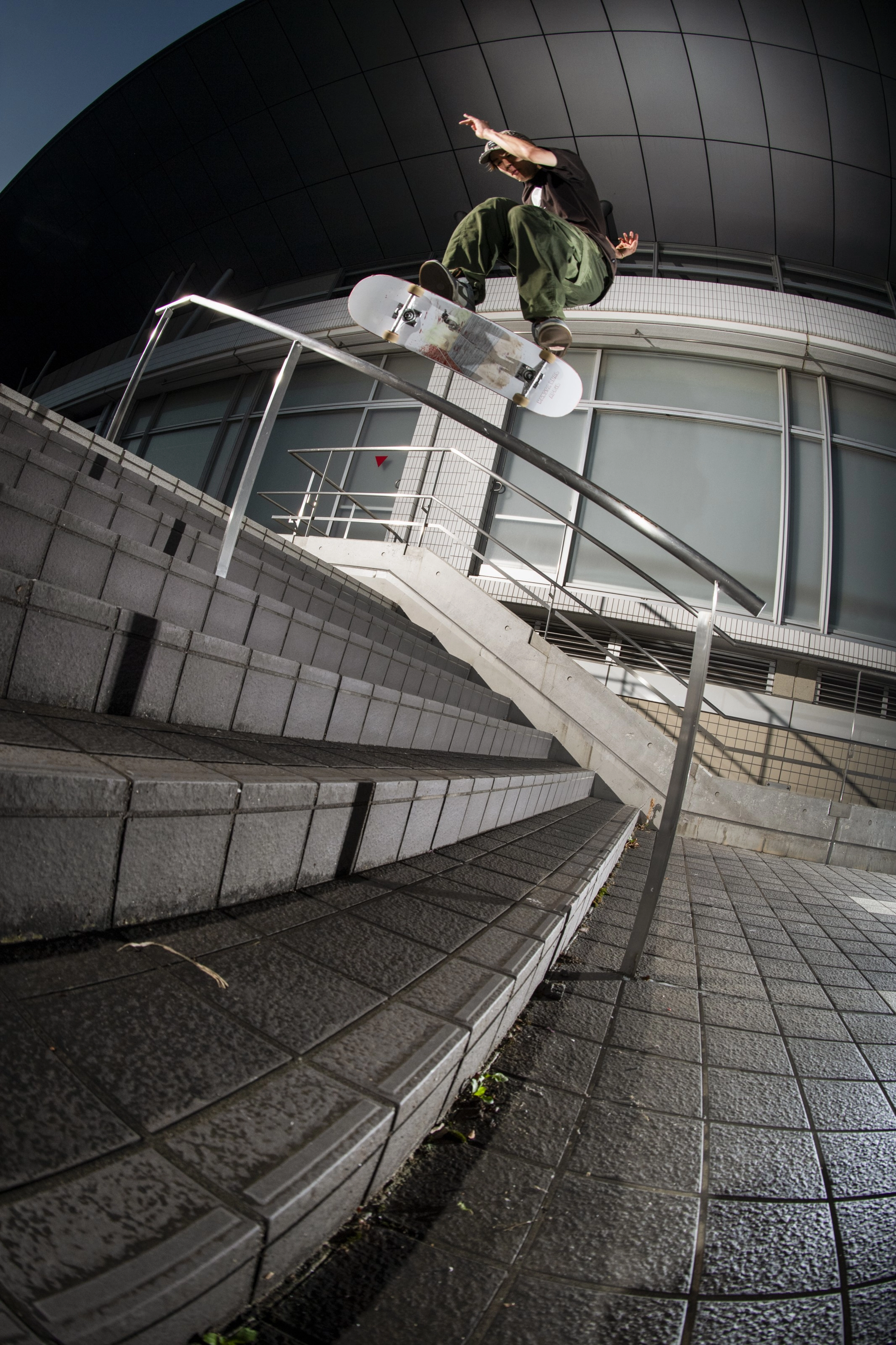 Yuto Horigome jumps a stair rail on his skateboard, wearing olive green pants and a black tee shirt. The concrete steps underneath are wet, and there is a building in the background. His trick is lit up in the dark, and there is a glimpse of the night sky.