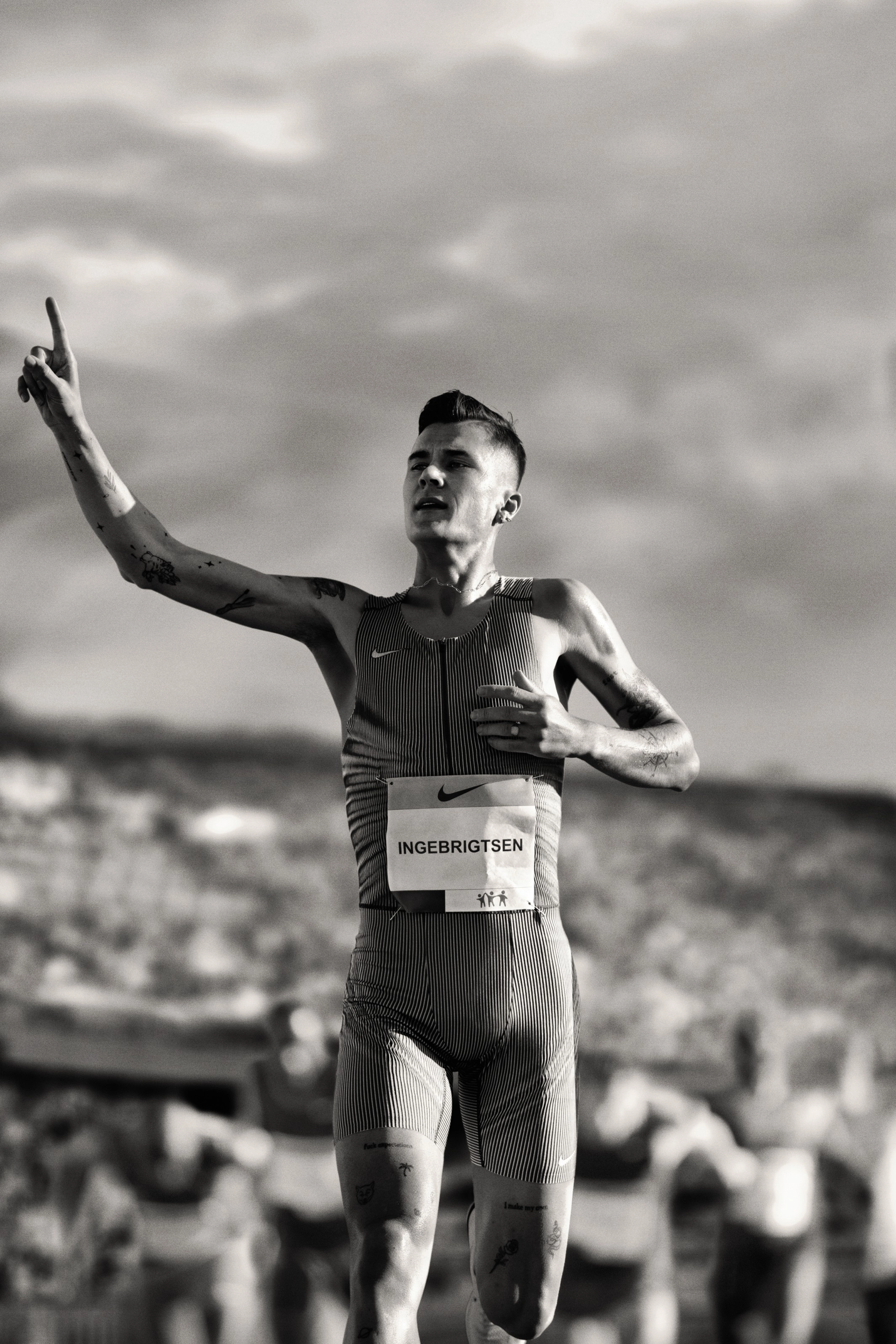 Black and white photo of Jacob Ingebrigtsen holding up his arm, celebrating his number 1 result. There is a crowd in the stadium behind him.