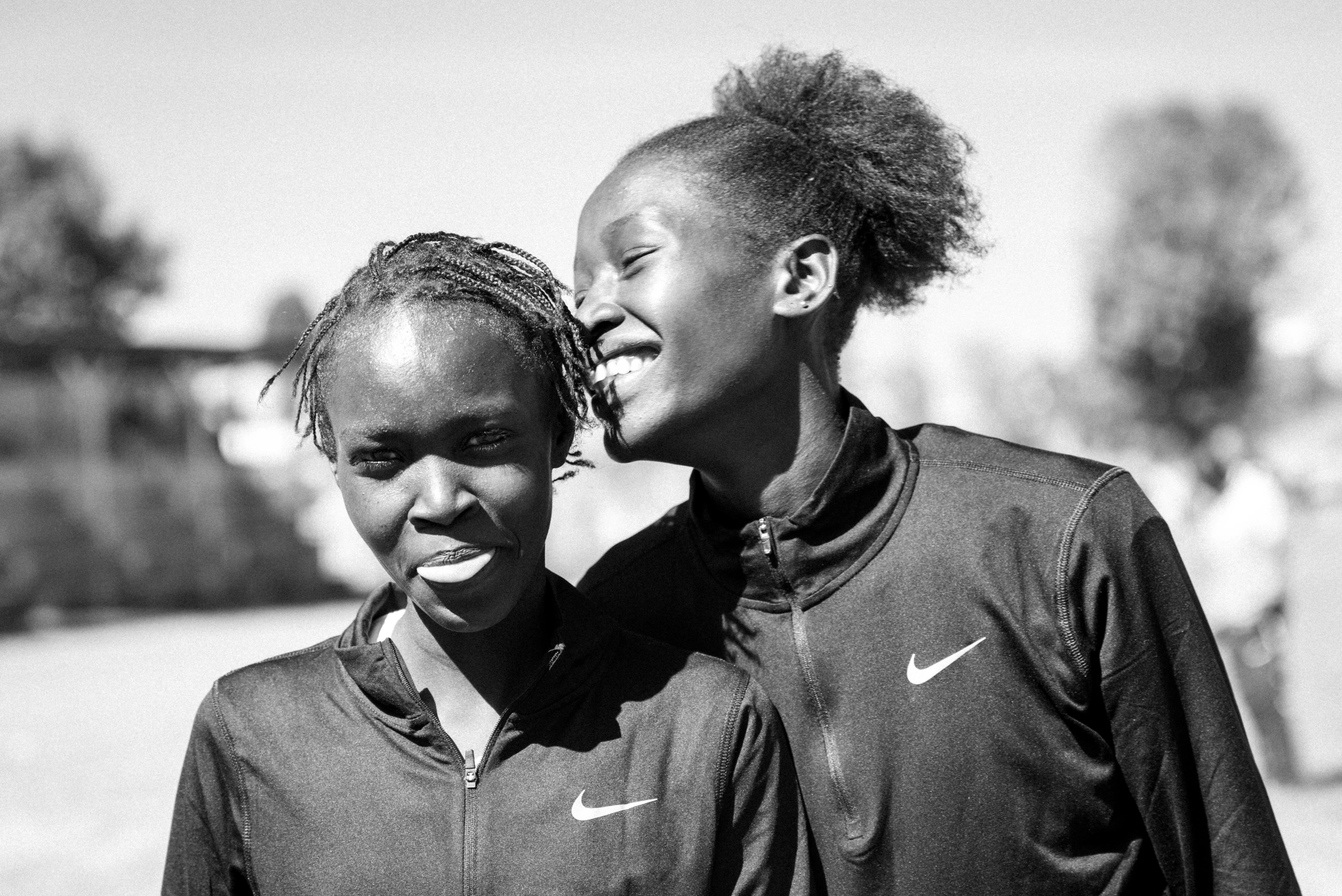 Black and white photo of two members of the Nala Track Club standing outdoors wearing Nike running jackets, smiling side by side each other.