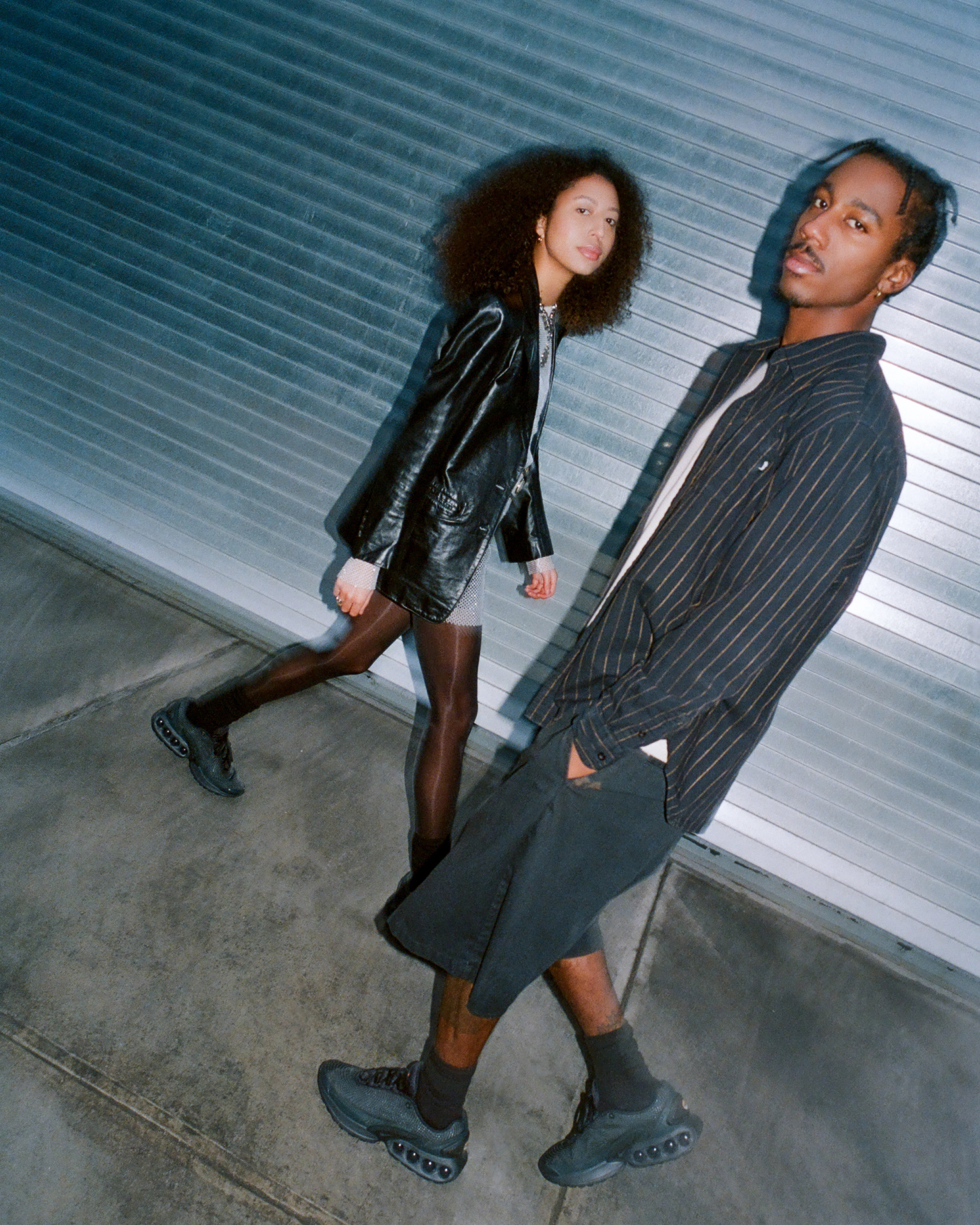 Two models, one male and the other female, wearing the Nike Air Max Dn in black while walking outside on the sidewalk with a metallic garage door in the back